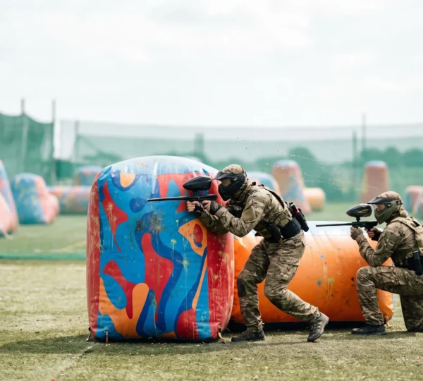 Duel tactique intense entre deux joueurs de speedball accroupis derrière des bunkers gonflables sur un terrain symétrique